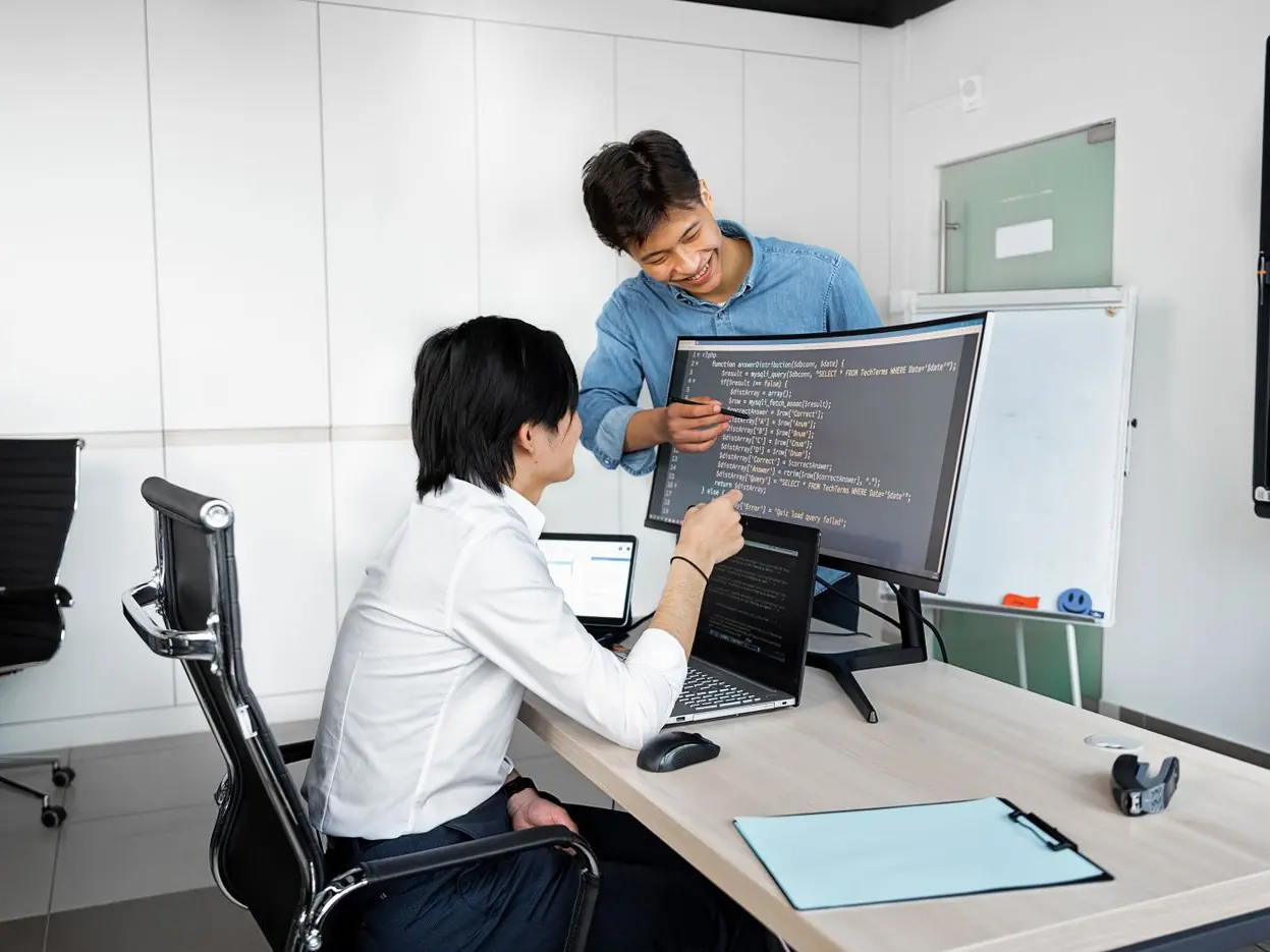 A young intern at an office computer while a colleague points out code on the screen